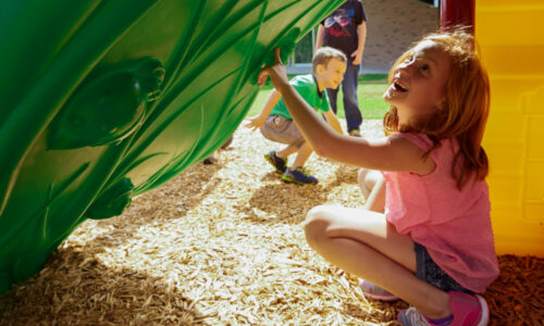 Children playing on a colorful playground structure, with a girl smiling while crouching near a green slide and a boy exploring in the background, highlighting safe and engaging outdoor play.